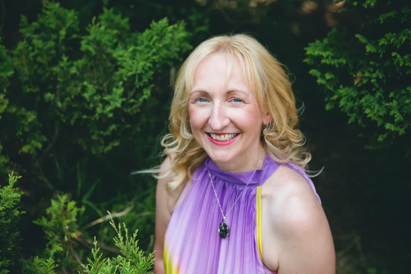 A smiling blonde woman with blue eyes wearing a purple dress and a necklace, framed by green foliage.