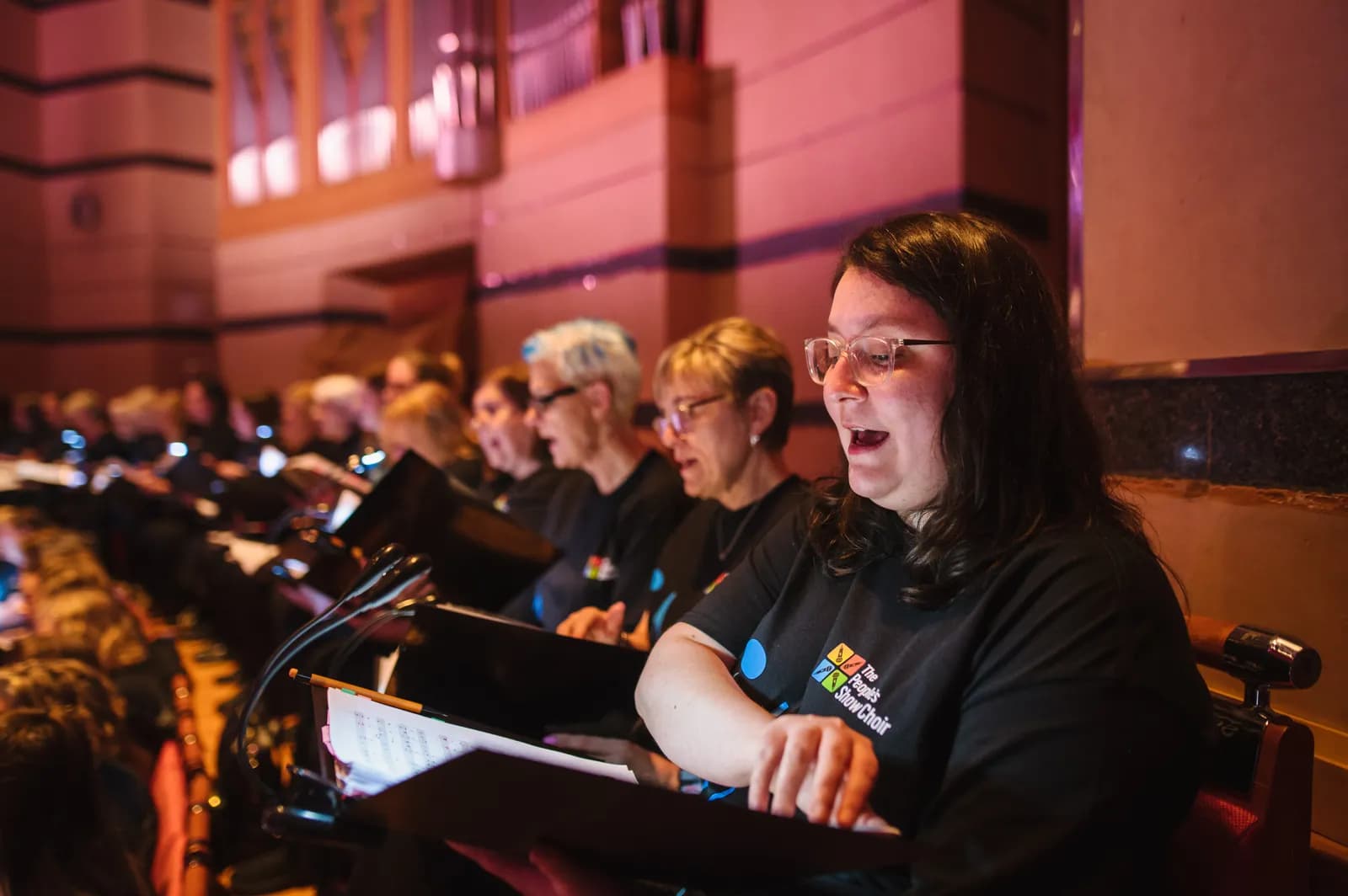 A woman in glasses and a black t-shirt with a colorful logo sings in a choir, with other singers visible behind her.